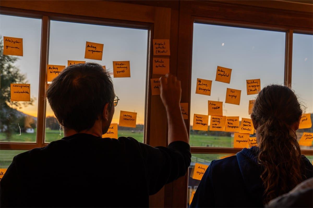 Beim Event Storming klebt ein Kollege eine Haftnotiz an die Fensterfront, eine Kollegin schaut zu