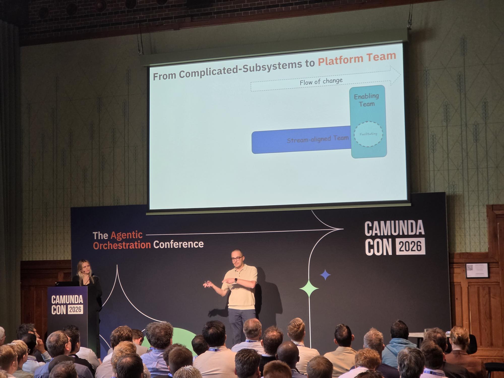 Maria Alish und Thomas Heinrichs auf der Bühne der CamundaCon 2026 vor der Slide "From Complicated-Subsystems to Platform Team".